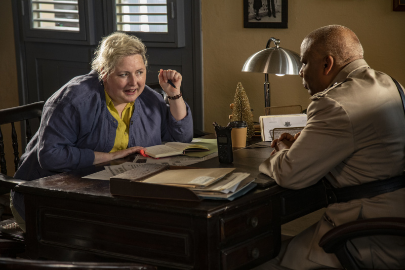  Jennifer ( Siobhán McSweeney ) interroge le commissaire Selwyn Patterson ( Don Warrington ) à son bureau dans son bureau. Elle tient un stylo et se penche en avant avec enthousiasme, et il y a un cahier devant elle.