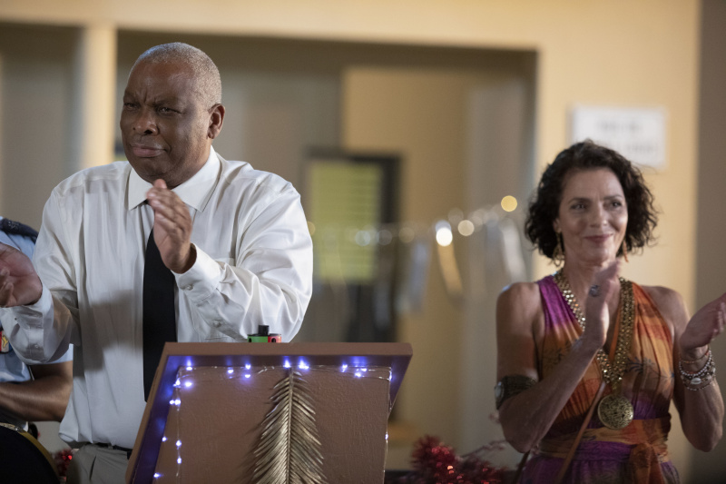  Selwyn (Don Warrington) și Catherine (Élizabeth Bourgine) aplaudă la aprinderea luminilor de Crăciun. Selwyn stă în spatele unui pupitru.