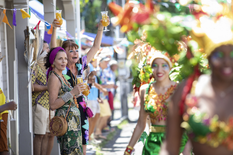  Un alambic din Death In Paradise's 2023 Christmas special showing Catherine (Élizabeth Bourgine) and Melanie (Doon Mackichan) attending a Christmas carnival. Melanie is clearly having a great time and holding two glasses of cocktails in the air.