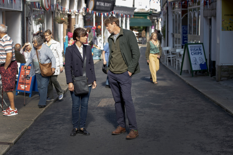   Un fotograma del episodio uno de Beyond Paradise que muestra a la DS Esther Williams (Zahra Ahmadi) y al DI Humphrey Goodman (Kris Marshall) parados en medio de Shipton Abbott.'s main shopping street. He has his hands in his pockets, and she is staring at him from beneath her fringe.