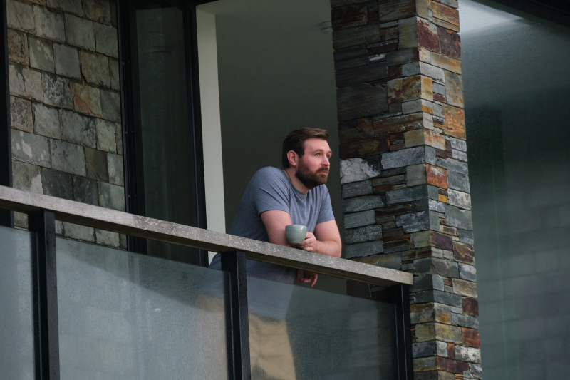  Miles (James McArdle) stoji na balkonu svoje impresivne, a stroge hiše, ki je zgrajena iz črne kovine in skrilaste opeke. Naslanja se na pregrado in nanjo odlaga skodelico čaja ter gleda na obzorje.