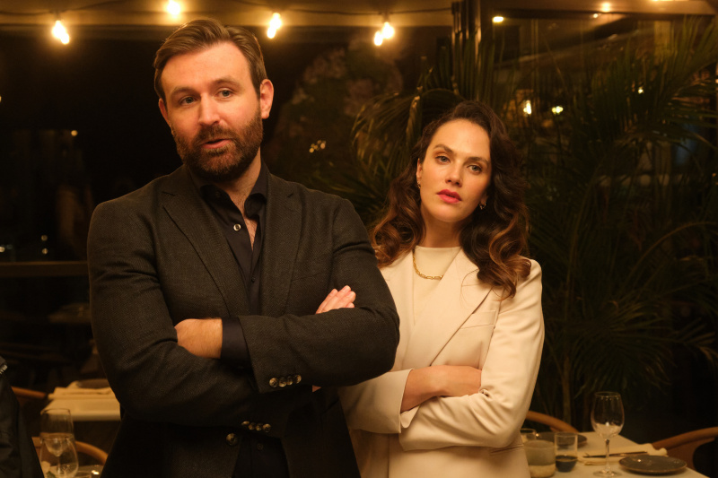  Miles (James McArdle) in Lucy (Jessica Brown Findlay) stojita v restavraciji s pravljičnimi lučmi, obešenimi po stropu. Oba nosita dragi obleki, Miles v črnem in Lucy v belem, oba imata prekrižane roke in nista videti navdušena.