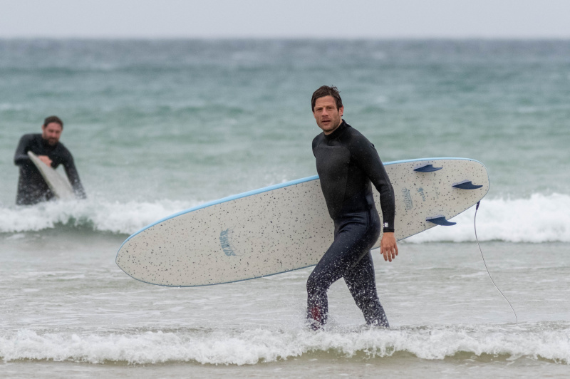  Pete (James Norton) stoji na robu morja v neoprenski obleki z desko za deskanje pod desno roko, medtem ko stopa proti plaži. V ozadju je tudi Miles (James McArdle) v neoprenski obleki in vzame svojo desko za deskanje.