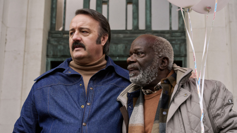   Tony Bubbins ในแจ็กเก็ตยีนส์ในบท Mammoth และ Joseph Marcell ในโค้ตสีเบจในบท Roger ใน Mammoth