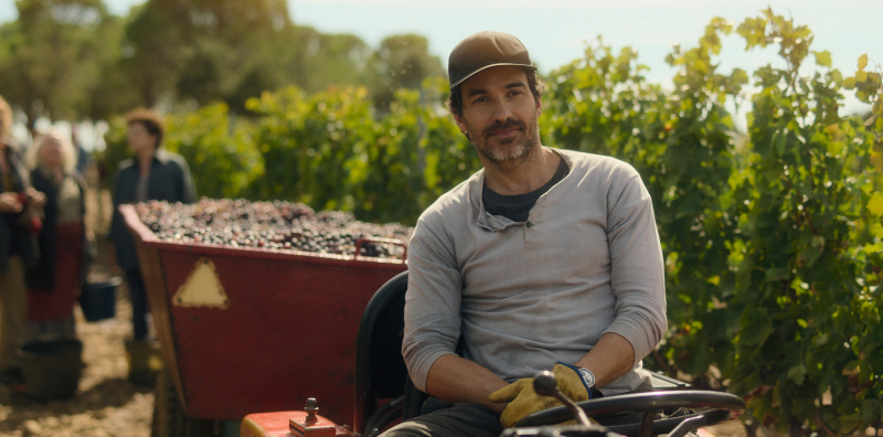   Amat (Santiago Cabrera) sitzt in einem Traktor mitten in einem Weinberg, hinter ihm ein Anhänger voller Weintrauben und im Hintergrund verschiedene Frauen, die Weintrauben pflücken