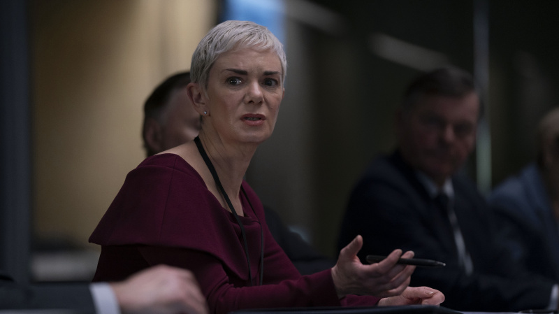   「COBRA: Rebellion」で、テーブルに座り動揺した表情を浮かべるアンナ・マーシャルを演じる、栗色のドレスを着たヴィクトリア・ハミルトン。