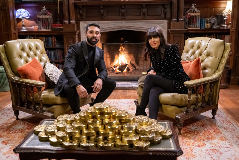   Jaz est assis dans un fauteuil du château des Traîtres en face de Claudia Winkleman dans un fauteuil séparé. Il y a un feu crépitant derrière eux, et devant la photo se trouve une table basse sur laquelle repose une pile tentaculaire de pièces d'or.
