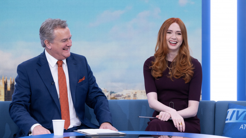   Hugh Bonneville în rolul lui Douglas într-un costum stă pe o canapea lângă Karen Gill, în timp ce Madeline într-o rochie maro pe fundalul londonez din Douglas este anulată
