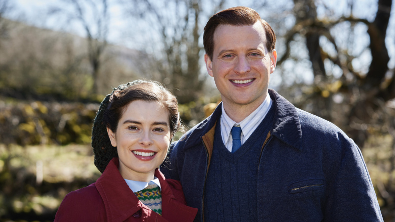  Rachel Shenton in einem roten Mantel als Helen und Nicholas Ralph in einem blauen Mantel als James stehen in „All Creatures Great and Small“ gemeinsam im Freien.