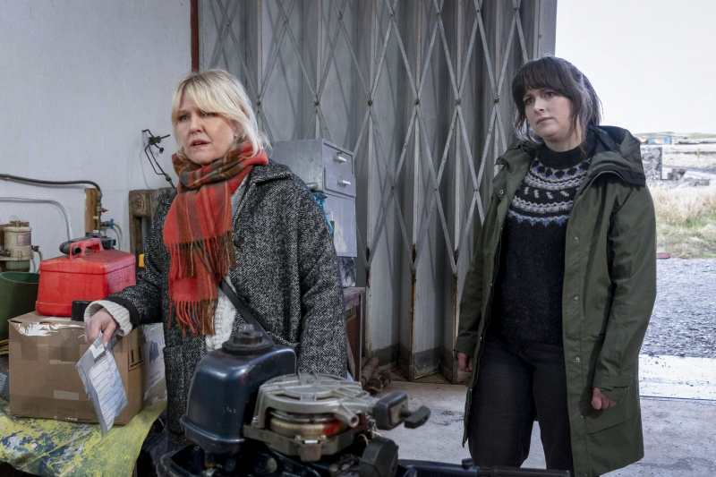  Calder (Ashley Jensen) y Tosh (Alison O.'Donnell) stand inside a warehouse or workshop of some kind - there is a large industrial concertina-style door behind them that's half open, and various items like a small bedside cabinet and an outboard motor scattered around in need of repair