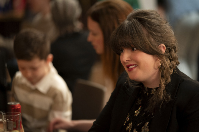  Tosh (Alison O)'Donnell) at a party with various people out of focus in the background, including a young boy. There are cans empty glasses and cans on the table in front of her. She is smiling a little uncomfortably.
