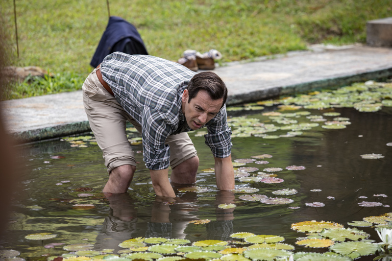   Neville (Ralf Little) se tient dans l'étang de la maison de retraite Paradise Bary, avec son pantalon retroussé, accroupi avec ses avant-bras pêchant dans l'eau.