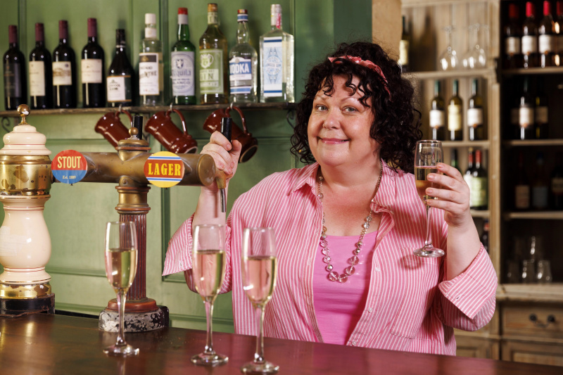  Gloria (Sue Vincent) se tient derrière la voiture à La Couronne, souriant à la caméra avec une main sur les escarpins et l'autre tenant un verre de champagne