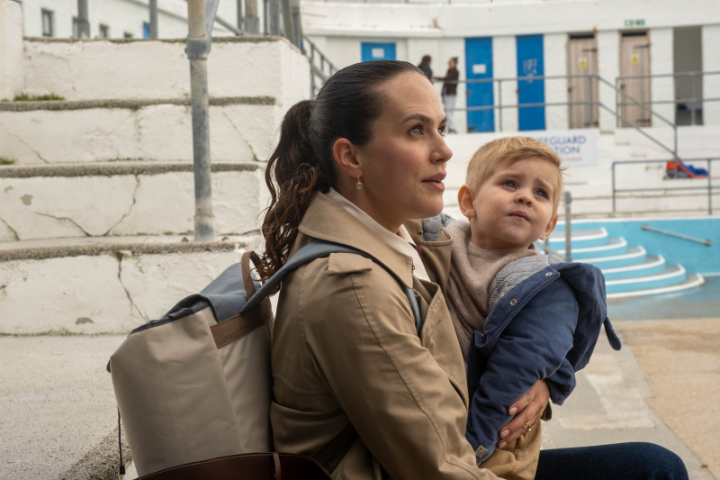  Lucy (Jessica Brown Findlay) está en un lido, lleva al bebé David en brazos y lleva una mochila grande. Parece estresada y ansiosa.