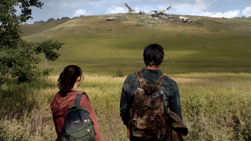  Ellie (Bella Ramsey) ja Joel (Pedro Pascal) seisavad seljaga kaamera poole ja vaatavad kauguses olevat künka, mille otsas on allakukkunud lennuki rusud.