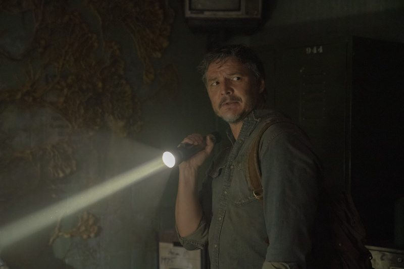  Pedro Pascal Joelina, kes seisab pimedas ruumis, kus tema ainus valgus tuleb tõrvikust's holding. He is turned towards the camera and looking apprehensive.