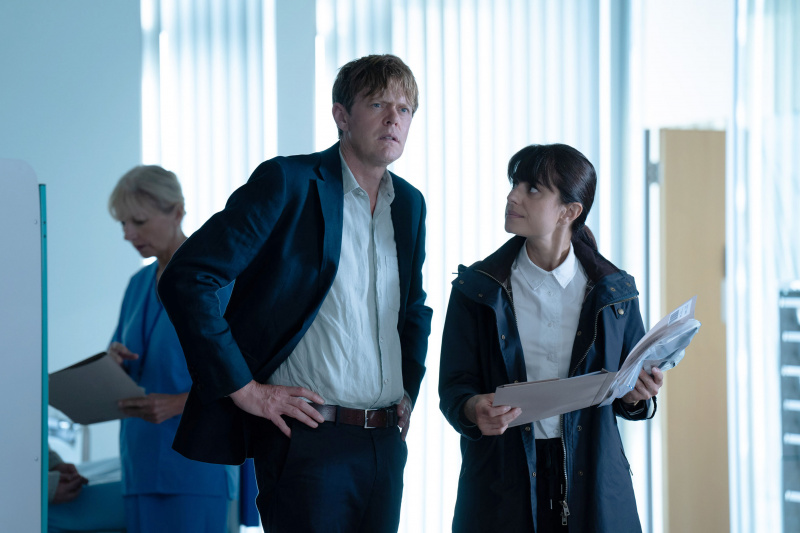 Beyond Paradise Staffel 2, Folge 5: DI Humphrey Goodman (Kris Marshall) und DS Esther Williams (Zahra Ahmadi) stehen auf einer Krankenstation. Er hat die Hände in die Hüften gestemmt und sie hält einen offenen Ordner mit medizinischen Notizen in der Hand.