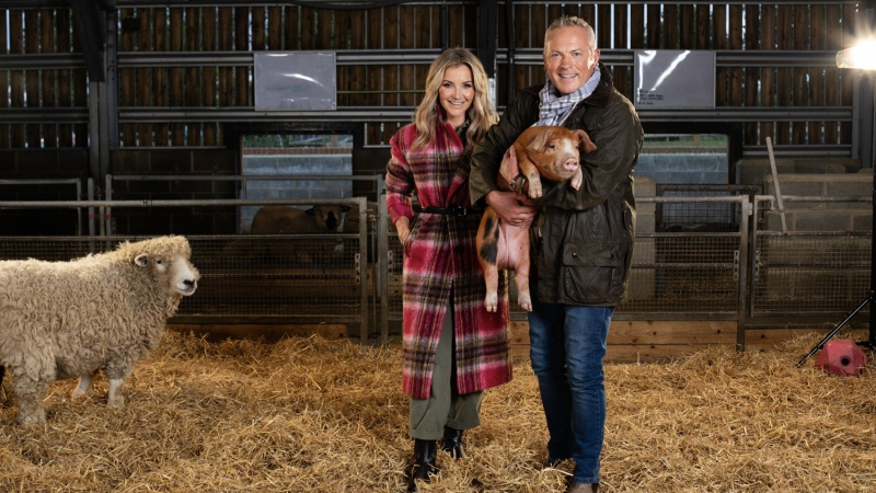   Helen Skelton y Jules Hudson en un granero con Jules sosteniendo un lechón y una oveja mirando en Winter on the Farm.