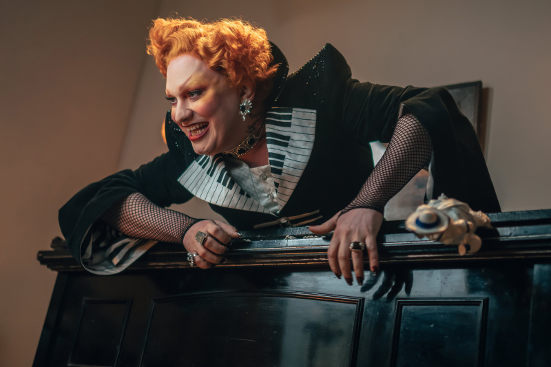  Ο μαέστρος (Jinkx Monsoon) σκαρφαλώνει μέσα από ένα πιάνο, στηρίζεται στο πλαίσιο και χαμογελάει απειλητικά, φορώντας ένα αστραφτερό μαύρο ρούχο με πέτα που μοιάζουν με πληκτρολόγιο πιάνου