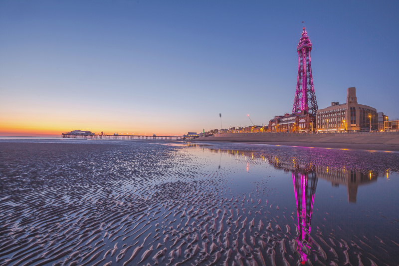  Näkymä Blackpoolin merenrannalle otettuna rannalta. Hiekka on märkää, ja horisontissa on kultainen auringonnousu. Blackpool Tower on valaistu magenta valolla.