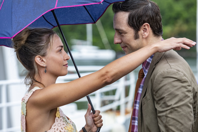 Chelsea Edge și Ralf Little în rolul lui Sophie și Neville în Death In Paradise. Se caută unul în celălalt's eyes and smiling. Sophie has one arm resting on Neville's shoulder, and is holding up an umbrella with her other hand.