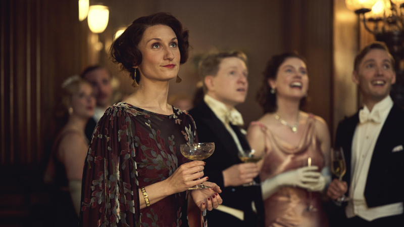   Bessie Carter tumedas lillelises kleidis, hoides klaasi, mängib Nancy Mitford filmis Outrageous