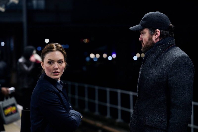   Strike: The Ink Black Heart – Robin (Holliday Grainger) și Strike (Tom Burke) stau în haine întunecate lângă râu.