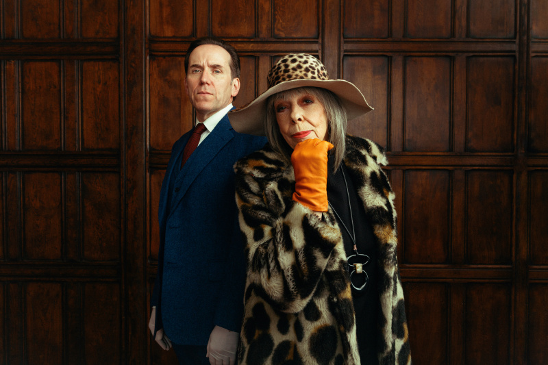  Professor Jasper Tempest (Ben Miller) und seine Mutter Adelaide (Frances de la Tour) stehen vor einer mit Mahagoni getäfelten Wand. Er trägt einen Anzug und weiße Plastikhandschuhe und steht aufrecht. Sie trägt einen großen Pelzmantel und einen breitkrempigen Hut mit Leopardenmuster, dazu orangefarbene Lederhandschuhe, und beugt sich vor, ihr Kinn ruht auf ihrer rechten Hand.