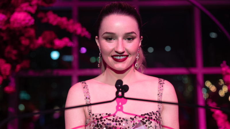   Kaitlyn Dever comme Belle Gibson souriant dans une robe à paillettes parlant dans un microphone.