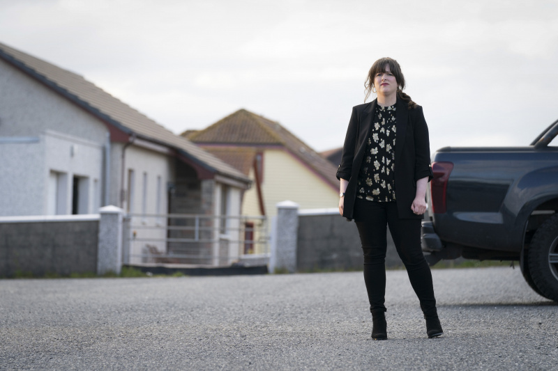  Tosh (Alison O'Donnell) stands in a driveway next to a pick-up truck. She looks a little hesitant. She is dressed up smarter than her usual police gear, in a black blazer and matching trousers, with a black and white floral top.