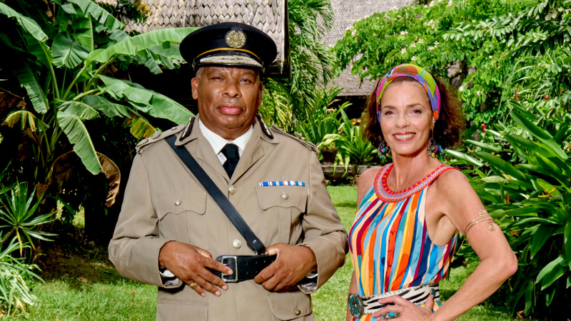   Bazı yemyeşil yeşilliklerin önünde Komiser Selwyn Patterson (Don Warrington) ve Catherine Bordey'in (élizabeth Bourgin) pozlanmış bir atış. Komiser işinde ve Catherine parlak çok renkli dikey şeritlere sahip bir elbise giyiyor.