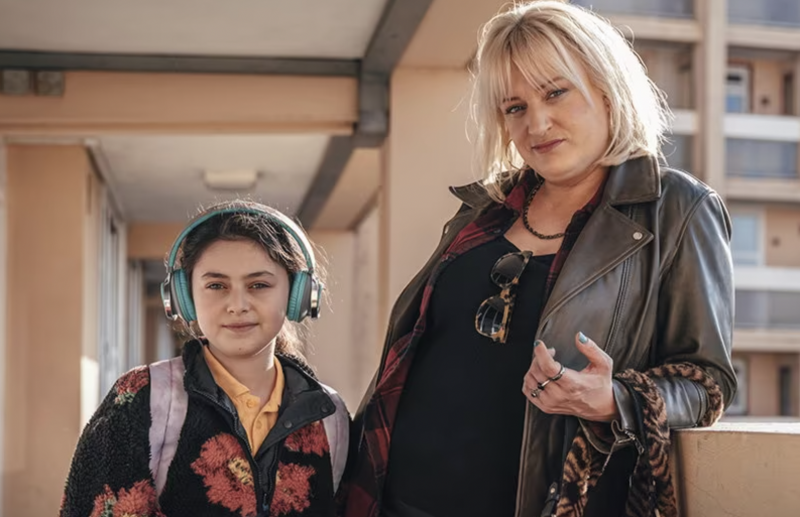   Fleur Tashjian so slúchadlami stojí vedľa Daisy May Cooper, ktorá drží kabelku