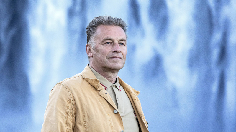   Chris Packham สวมแจ็กเก็ตมัสตาร์ดยืนอยู่หน้าน้ำตกในโลก