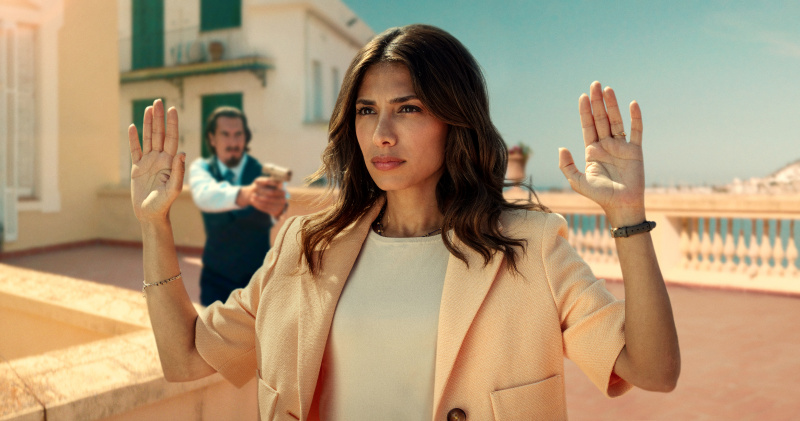   Un episodio di Still da Who Is Erin Carter? mostra Erin (Evin Ahmad) in piedi su un tetto, con le mani alzate mentre un uomo dietro di lei le punta una pistola contro