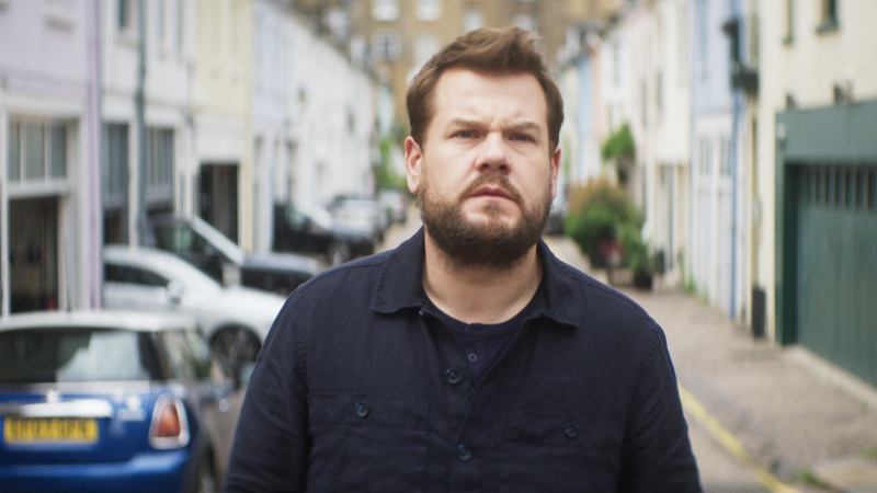   James Corden ในเสื้อสีเข้มดูหม่นหมองยืนอยู่บนถนนใน Mammals