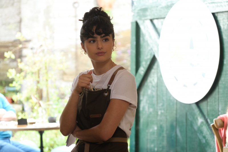   Zoe (Melina Sinadinou) ยืนอยู่ที่ทางเข้าประตูของ Ten Mile Kitchen โดยไม่สนใจที่จะจิ้มดินสอที่ด้านหน้าผ้ากันเปื้อนของเธอ