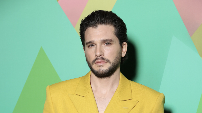   Kit Harington, yeşil bir arka planın önünde sarı ceketli.