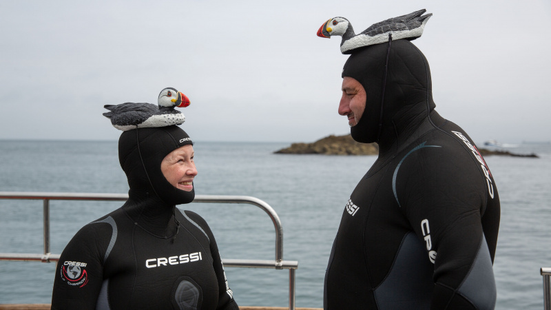   Wynne e Joanna in piedi su una barca in muta, entrambi con pulcinelle di mare di plastica legate alla testa