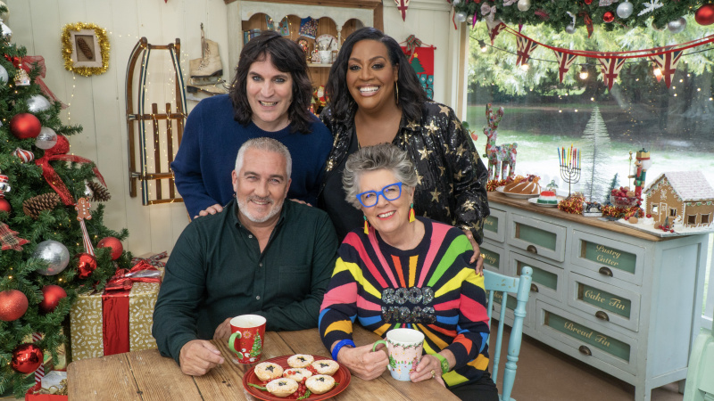   Paul Hollywood ในเสื้อเชิ้ตสีเข้มนั่งกับ Dame Prue Leith ในเสื้อจัมเปอร์สีรุ้ง และ Noel Fielding ในเสื้อจัมเปอร์สีเข้ม และ Alison Hammond ในเสื้อเบลาส์ที่เต็มไปด้วยดวงดาวยืนอยู่ด้านหลังพวกเขาในเต็นท์ซึ่งได้รับการตกแต่งสำหรับคริสต์มาสใน The Great British Bake Off