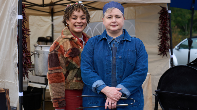 Rochenda Sandall v hnedom kabáne ako Fi a Siobhán McSweeney v modrom klobúku a kabát, keď Della stojí pred stanom v Amandalande.