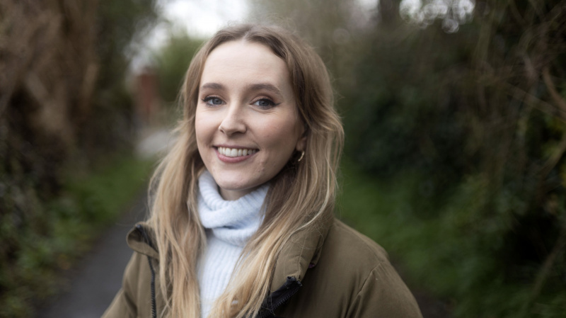   Rose Ayling-Ellis ในเสื้อโค้ทสีน้ำตาลและเสื้อสีขาวยืนอยู่ข้างนอกใน Signs for Change