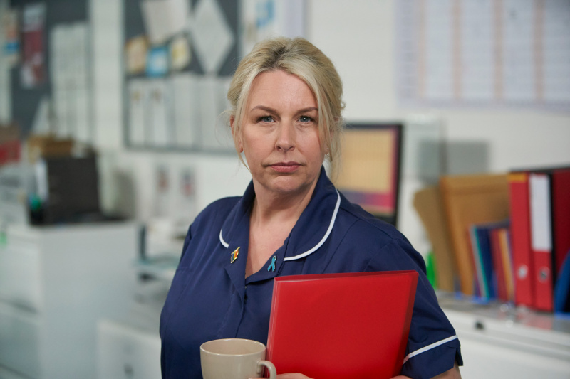   Hannah Walters เป็น Beth Relph สวมพยาบาลสีน้ำเงิน's uniform and holding a red clipboard
