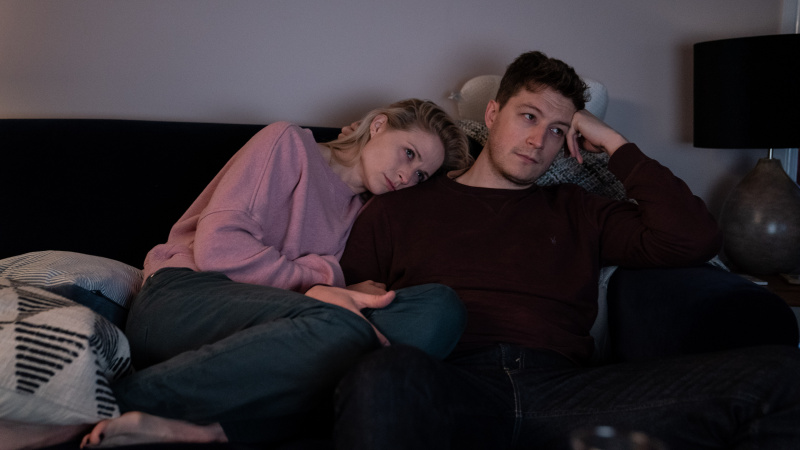   NIAMH ALGAR รับบทเป็น Dr. Lucinda Edwards และ LORNE MACFADYEN รับบทเป็น Tom Edwards ลูซินด้า's head is resting on Tom's shoulder as they sit together on the sofa