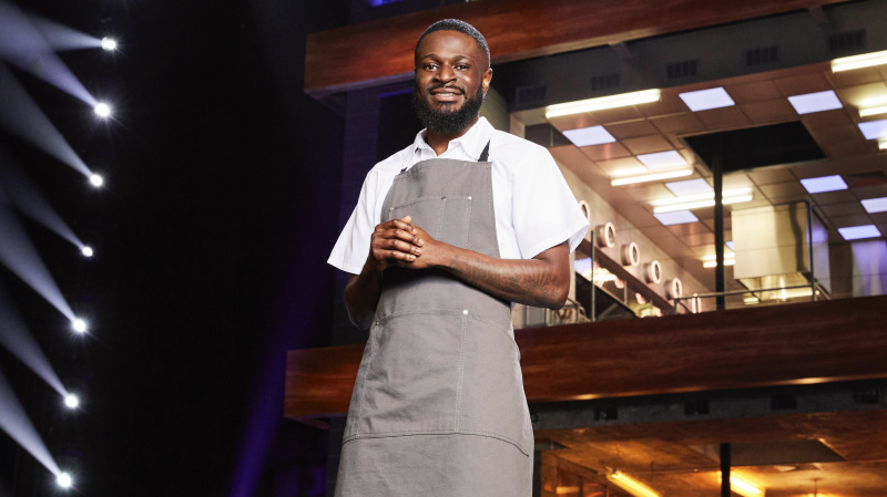   ผู้เข้าแข่งขัน Temi สวมเสื้อสีขาวและผ้ากันเปื้อนสีเทาหน้าห้องครัวใน Next Level Chef