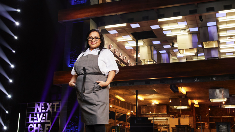  ผู้เข้าแข่งขัน Tia สวมเสื้อสีขาวและผ้ากันเปื้อนสีเทาหน้าครัวใน Next Level Chef
