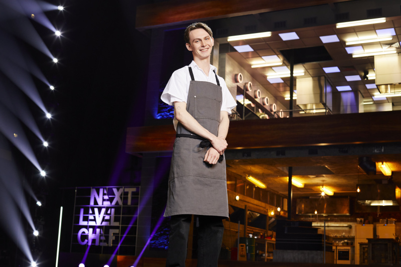   ผู้เข้าแข่งขัน Toby สวมเสื้อสีขาวและผ้ากันเปื้อนสีเทาหน้าห้องครัวใน Next Level Chef