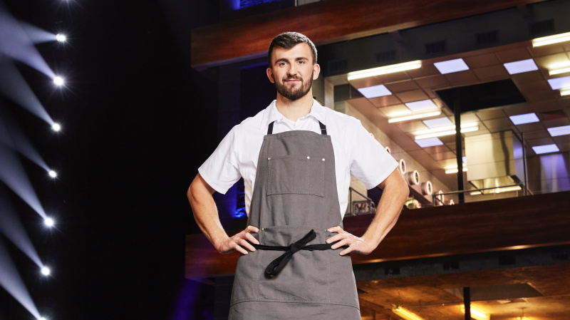   ผู้เข้าแข่งขัน Callum สวมเสื้อสีขาวและผ้ากันเปื้อนสีเทาหน้าห้องครัวใน Next Level Chef