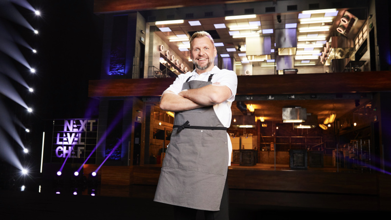   ผู้เข้าแข่งขันโกลด์สวมเสื้อสีขาวและผ้ากันเปื้อนสีเทาหน้าห้องครัวใน Next Level Chef
