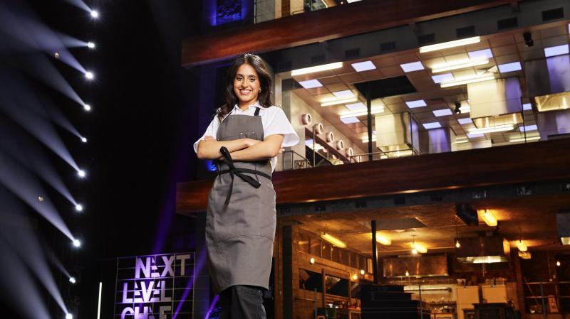   Gurpreet ผู้เข้าแข่งขันสวมเสื้อสีขาวและผ้ากันเปื้อนสีเทาหน้าห้องครัวใน Next Level Chef