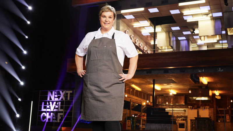   ผู้เข้าประกวด Kelly สวมเสื้อสีขาวและผ้ากันเปื้อนสีเทาหน้าห้องครัวใน Next Level Chef